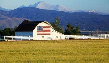 A barn with the USA flag painted on the side is flanked by farm fields and mountains.